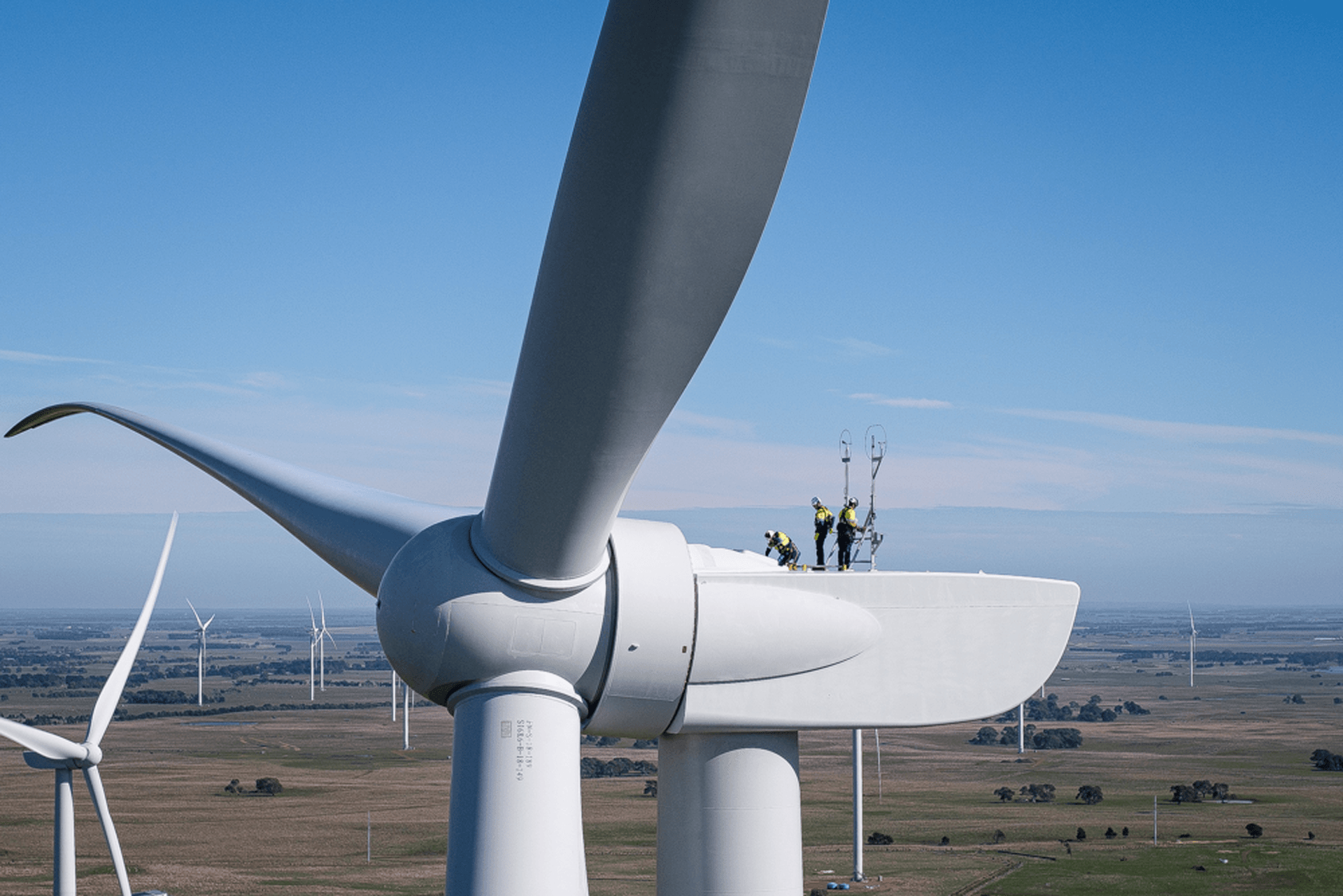 Three people standing on a wind turbine with a wind farm in the distance.